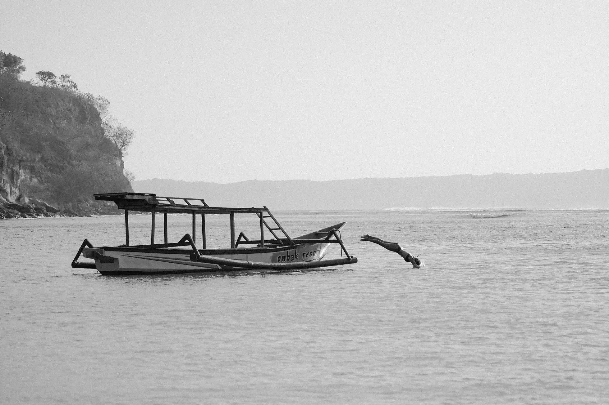 Somewhere Lombok, Bali, Indonesia — Traditional wooden boat anchored in calm waters with dramatic cliff backdrop, black and white coastal photography capturing serene maritime atmosphere.