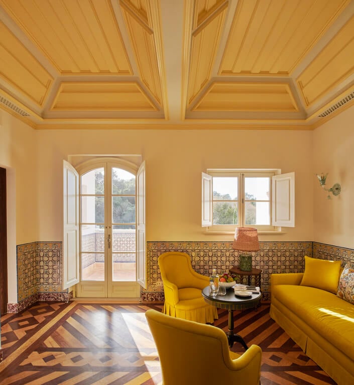 Vermelho, Europe — Sunlit sitting room with coffered wood ceiling, ochre walls, geometric tile wainscoting, mustard upholstered chairs, and terracotta patterned floor.