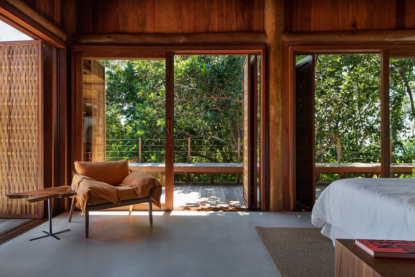Barracuda Hotel & Villas, South America — Teak bedroom suite with cognac leather chair, floor-to-ceiling glass doors opening to jungle canopy deck with metal railings.
