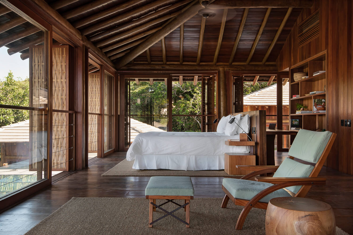 Barracuda Hotel & Villas, South America — Teak bedroom pavilion with glazed doors opening to forest views, white bedding, mid-century lounge chairs, exposed beam ceiling.