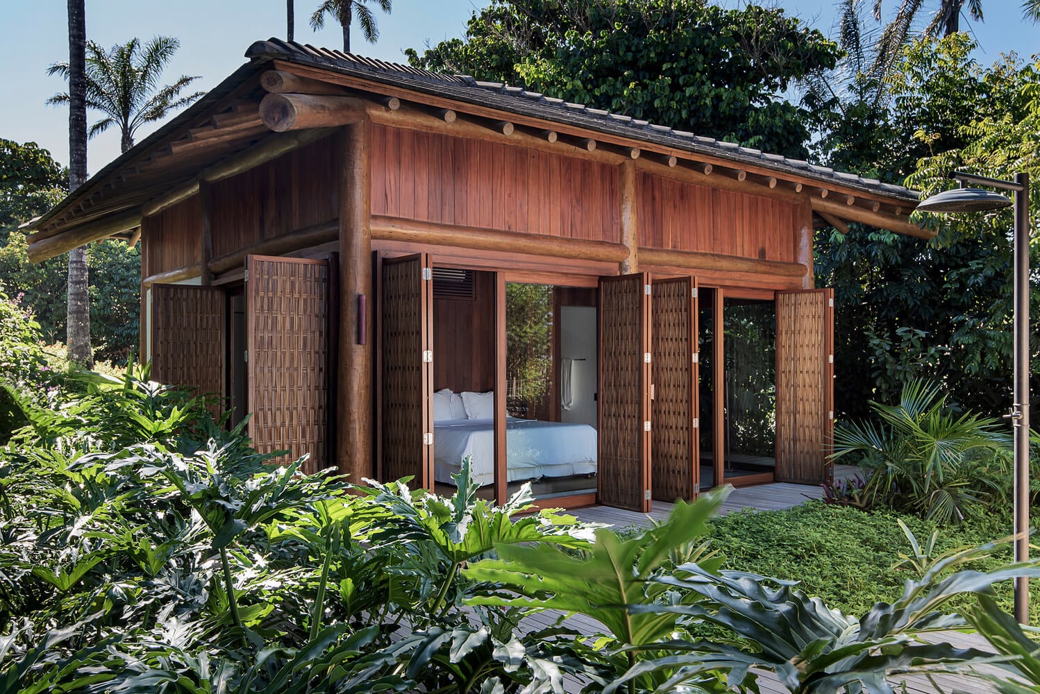 Barracuda Hotel & Villas, South America — Tropical villa with dark wood architecture, sliding glass doors, white bedding visible inside, lush green vegetation, afternoon light.