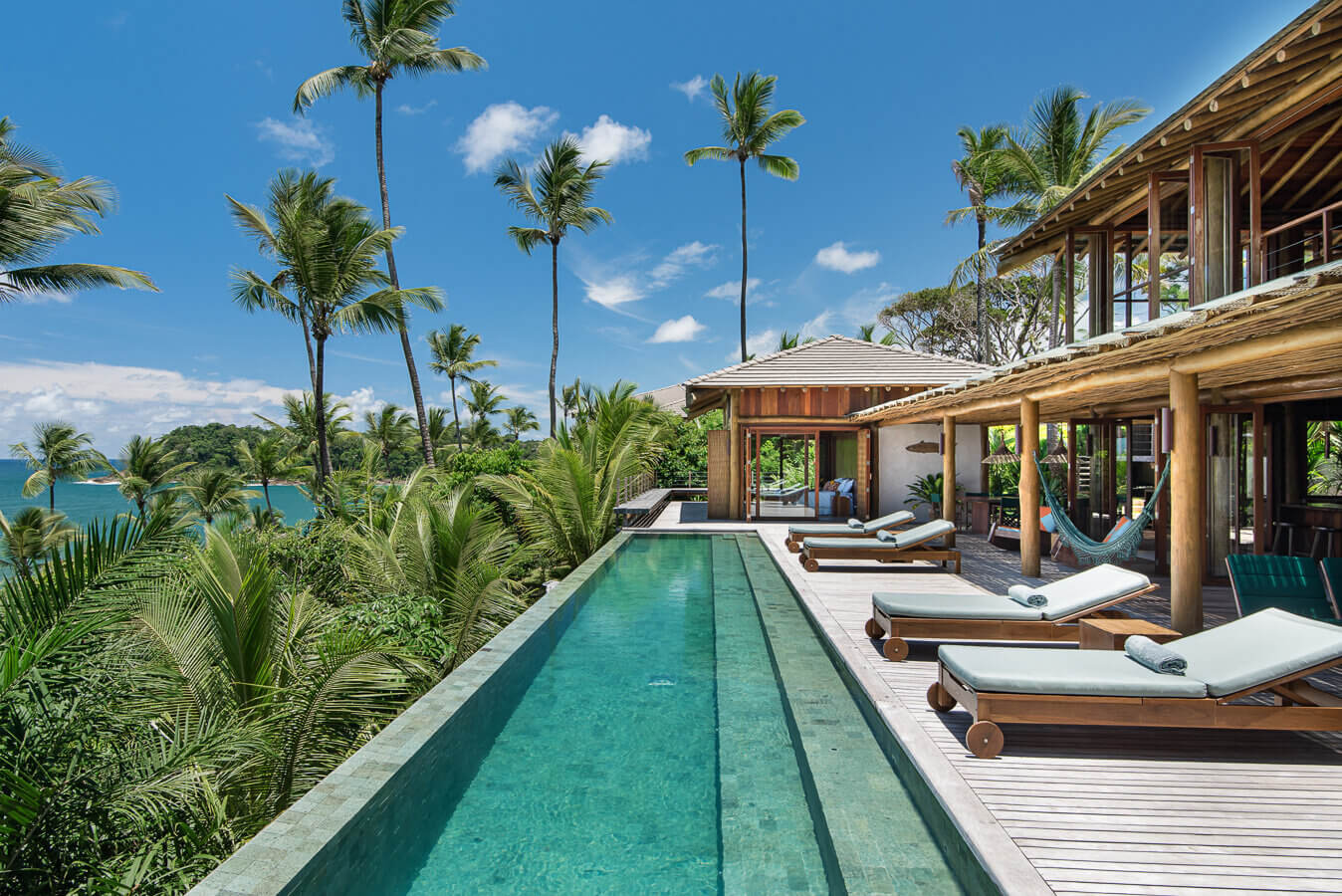 Barracuda Hotel & Villas, South America — Infinity pool with teak loungers overlooking tropical coastline, modern wooden pavilion with ocean views and palm trees.
