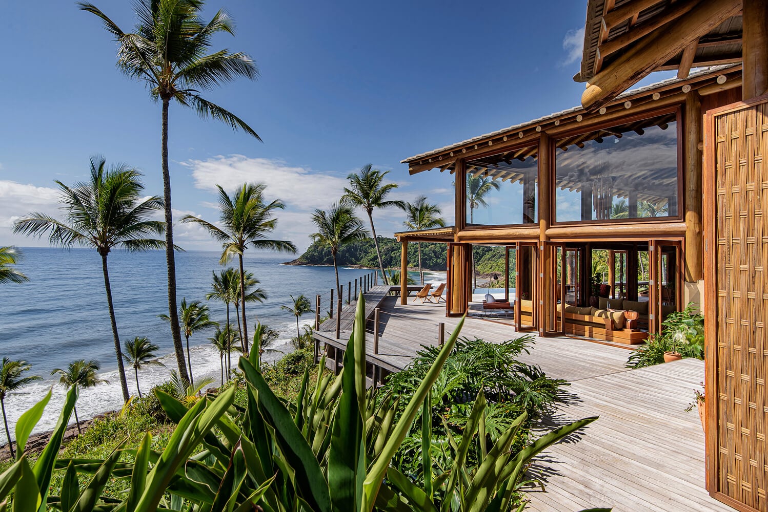 Barracuda Hotel & Villas, South America — Teak pavilion with expansive glazing overlooking palm-lined coastline and volcanic headland, warm wood decking and contemporary tropical architecture.