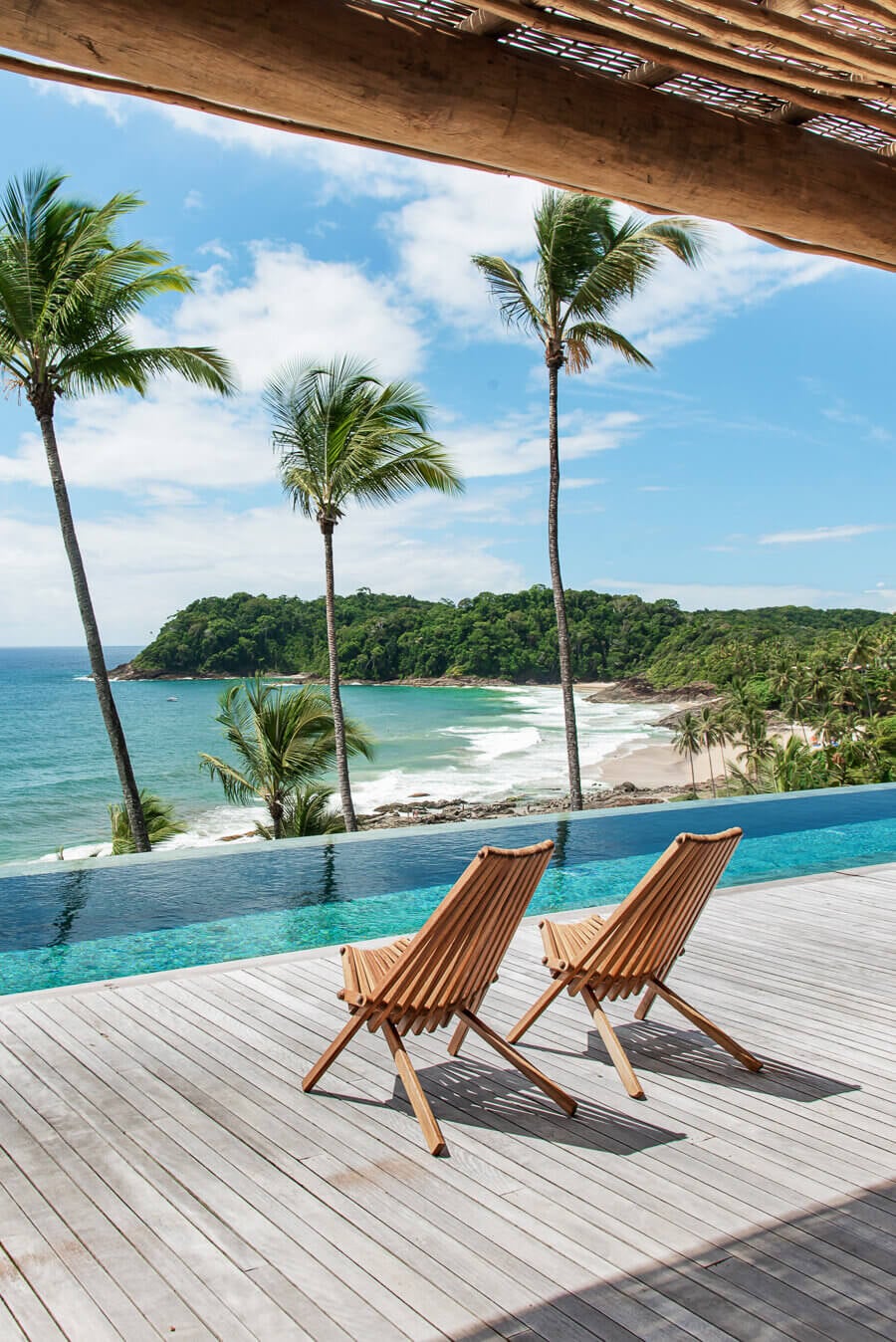 Barracuda Hotel & Villas, South America — Wooden deck with striped lounge chairs overlooking infinity pool, palm trees, and turquoise coastline with forested headland.