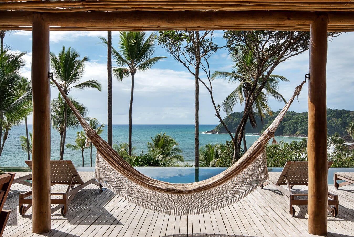 Barracuda Hotel & Villas, South America — Woven hammock suspended between timber posts on oceanfront deck overlooking turquoise waters and palm trees.