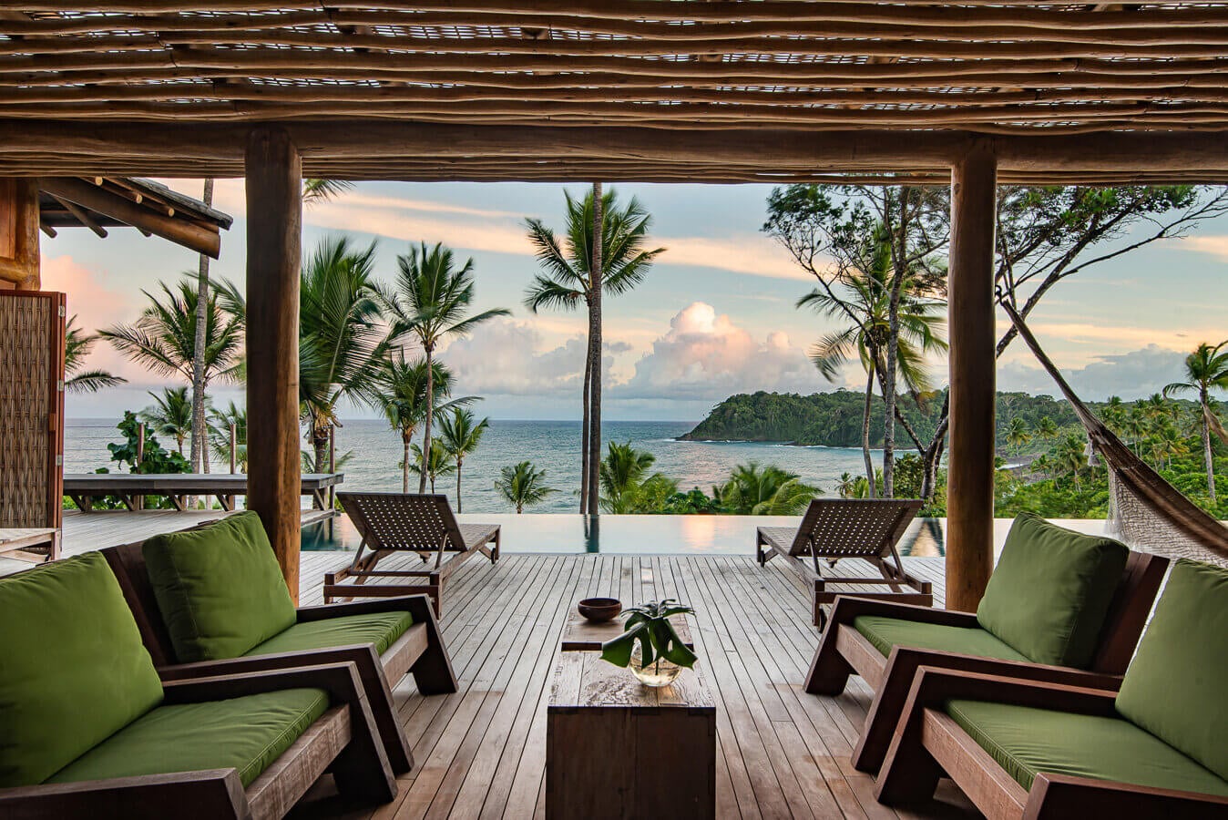 Barracuda Hotel & Villas, South America — Covered terrace with lime cushioned seating, teak loungers, ocean view, palm trees, wooden deck, tropical coast at sunset.
