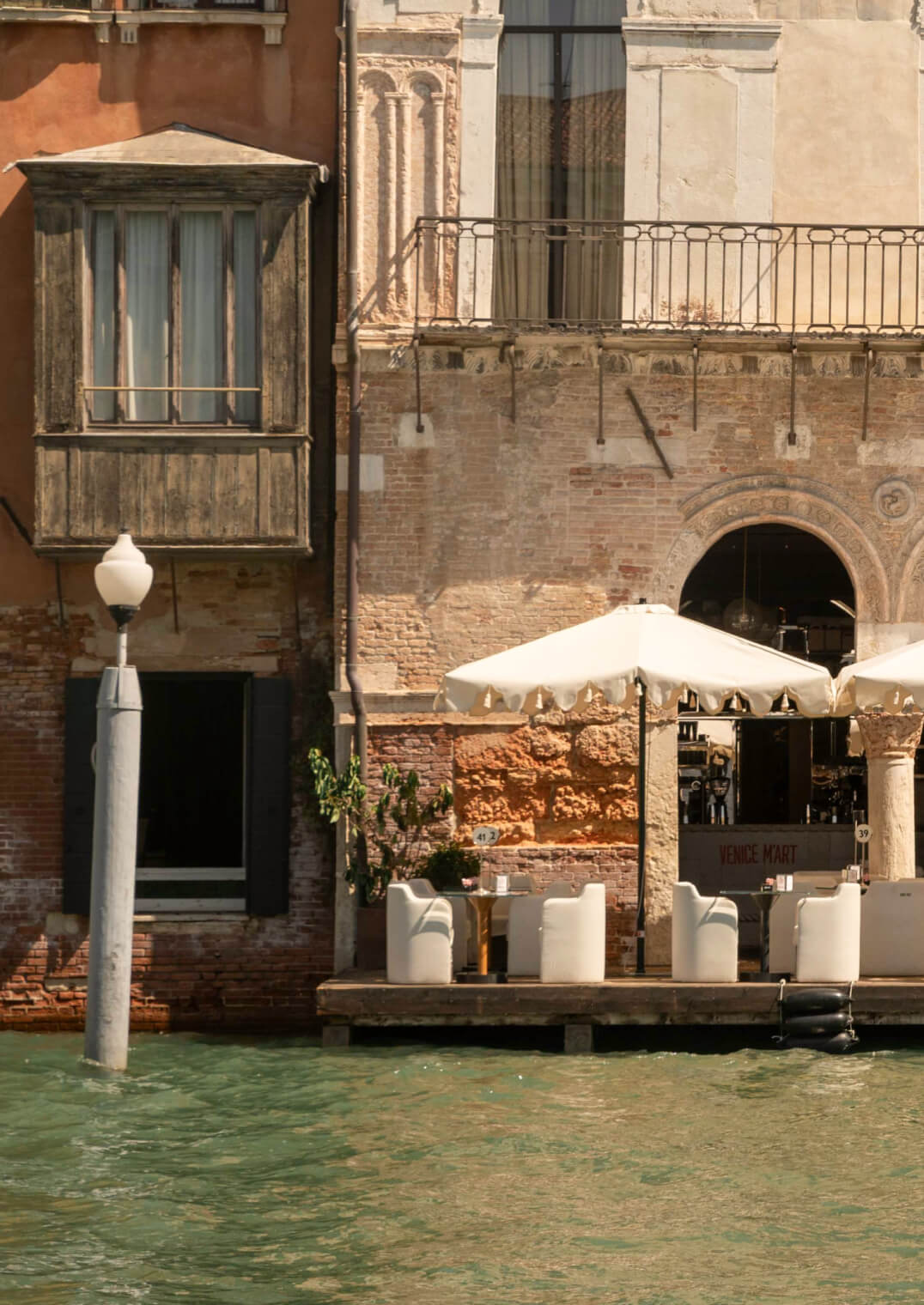 The Venice Venice Hotel, Venice, Italy — Waterfront terrace with white umbrellas, cream seating, and ochre brick architecture along the Grand Canal, noon light.