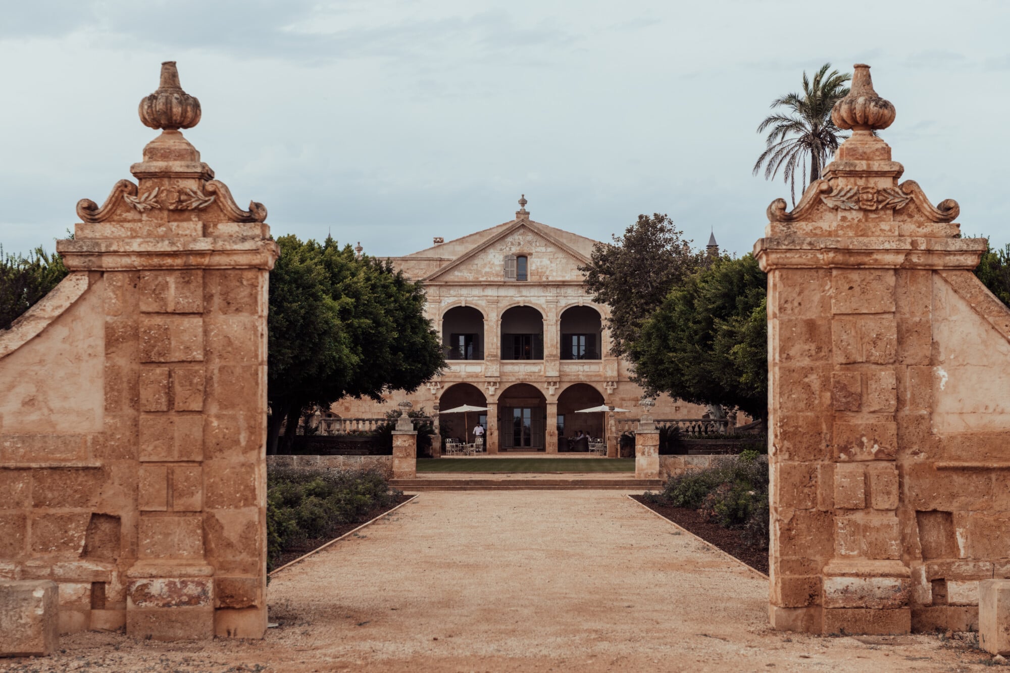 Son Vell, Balearic Islands — Terracotta stone gateposts with ornamental finials flanking a gravel drive toward a russet-hued manor house with arched loggias and pitched roof.