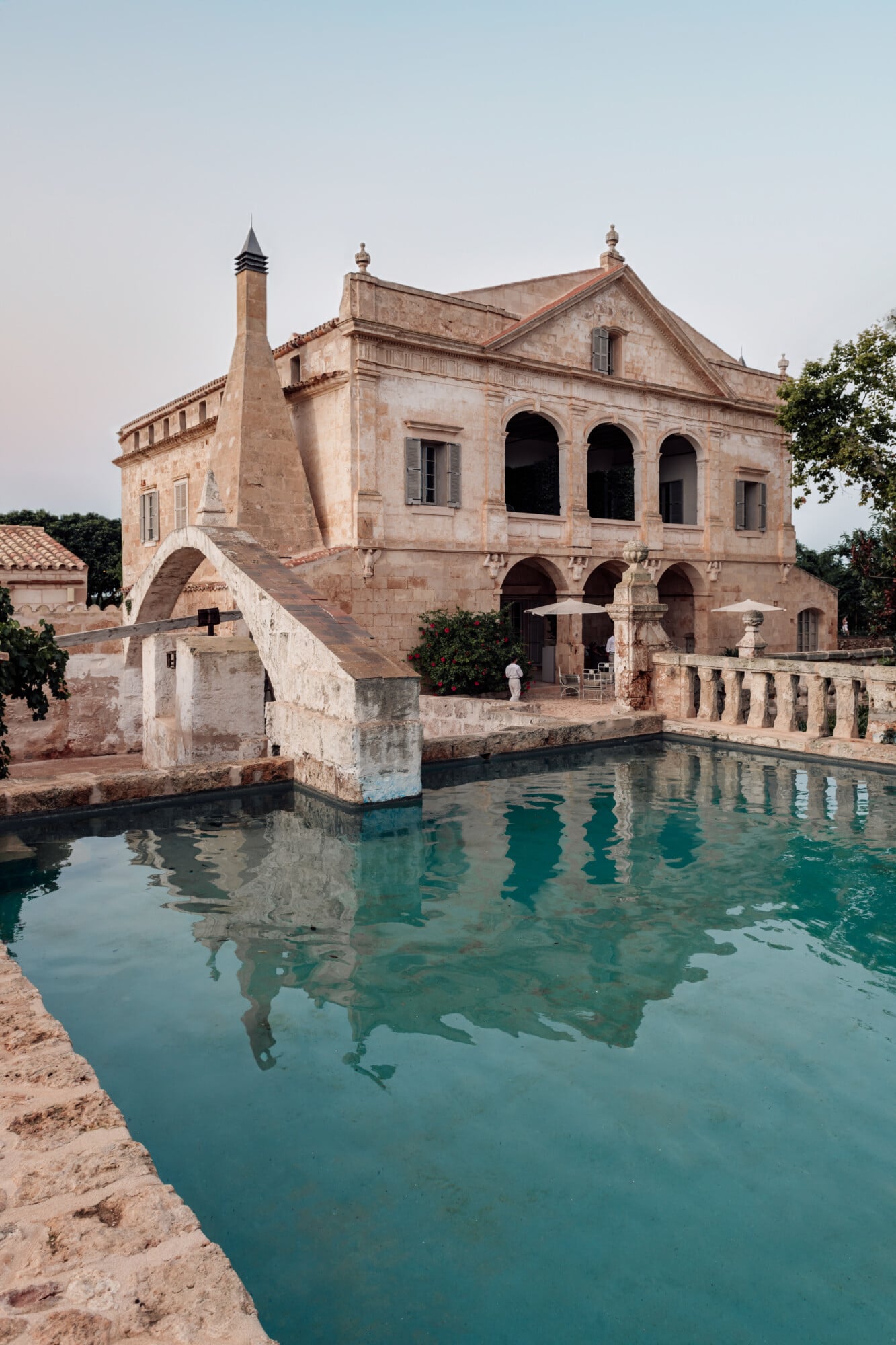 Son Vell, Balearic Islands, Spain — Ochre stone villa with arched loggia beside turquoise spring pool, Mediterranean architecture reflected in still water.