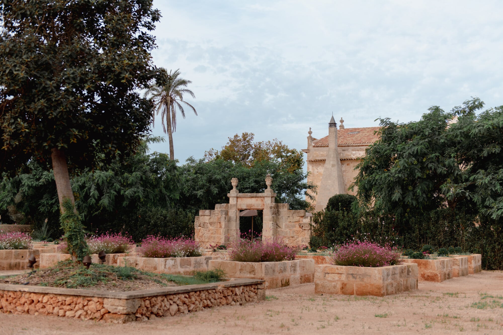 Son Vell, Balearic Islands, Spain — Ochre stone gatehouse with decorative urns framed by flowering shrubs and mature palms in Mediterranean garden courtyard.