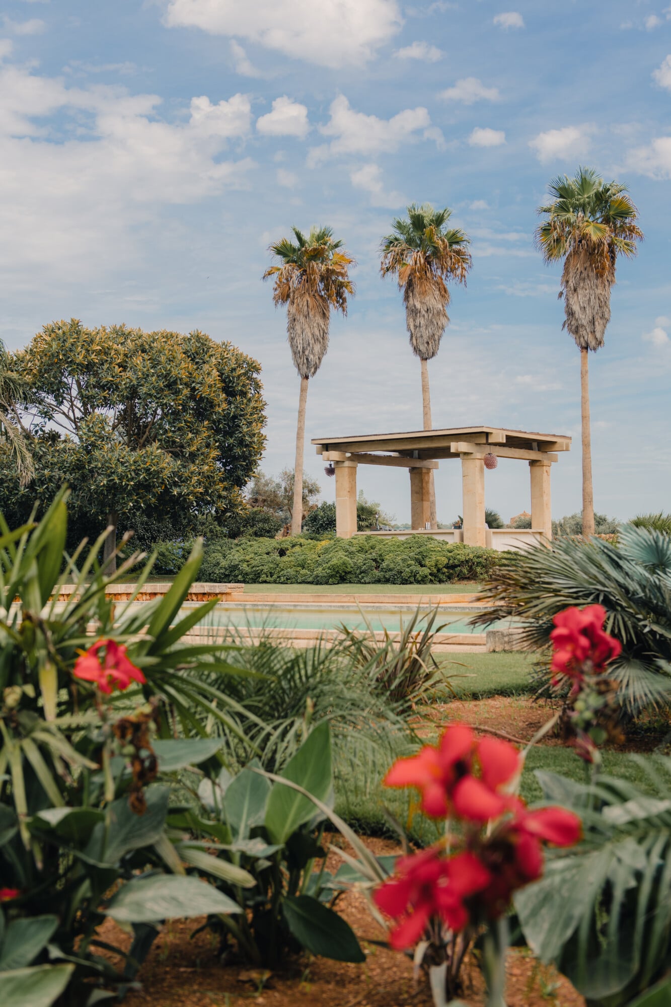 Son Vell, Balearic Islands — Poolside pavilion with pale stone columns and three tall palms, framed by red flowering plants and manicured gardens under blue sky.