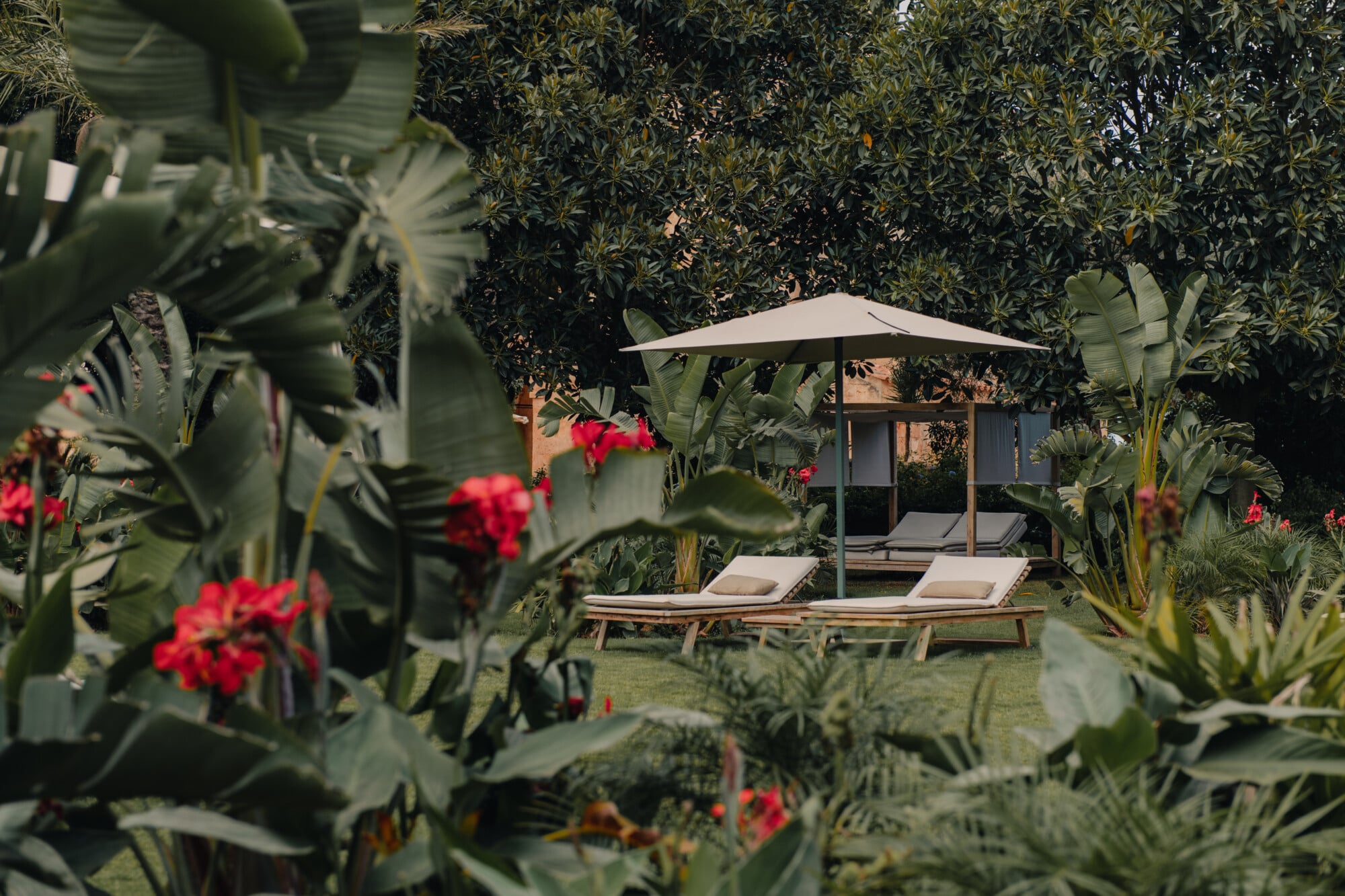 Son Vell, Balearic Islands — Shaded lounge chairs beneath umbrella in lush garden with red flowers, succulents, and Mediterranean vegetation framing relaxation space.