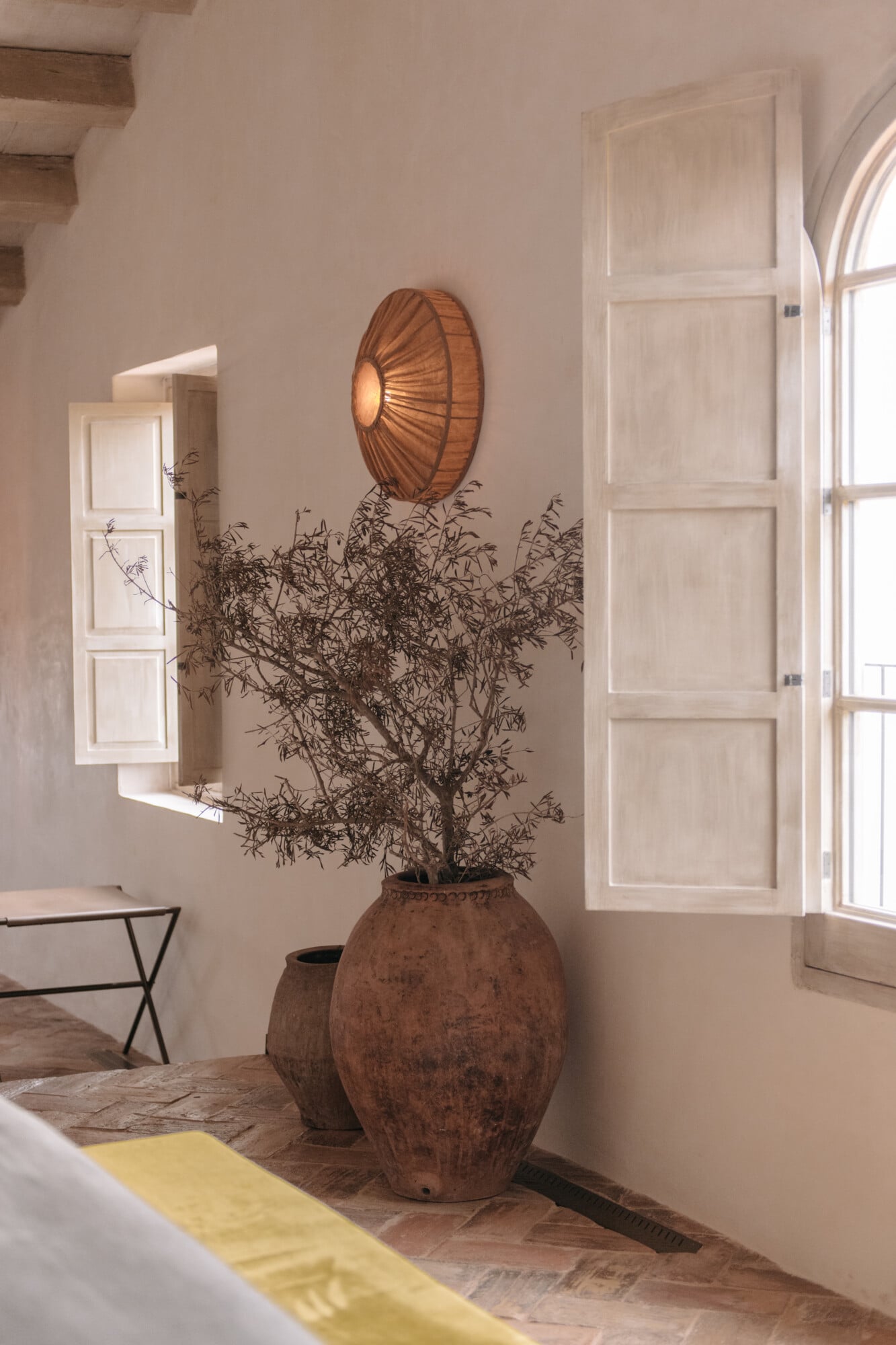 Son Vell, Balearic Islands — Minimalist room with dried olive tree in terracotta vessel, wooden pendant light, white shutters, and natural Mediterranean light.