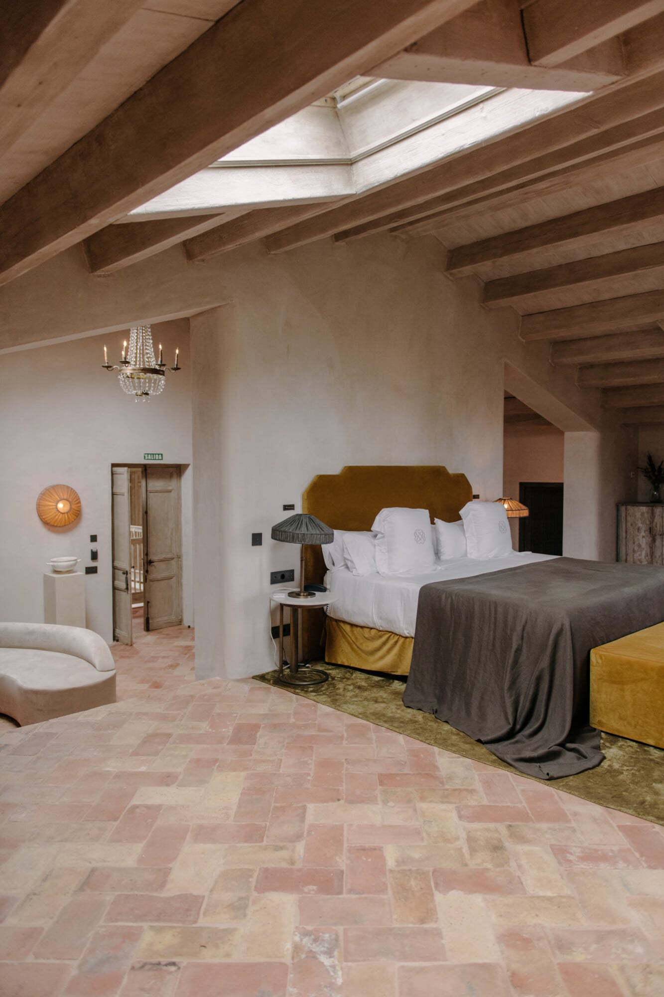 Son Vell, Balearic Islands, Spain — Attic bedroom with ochre headboard, exposed wooden beams, skylight, terracotta tile floors, and minimalist furnishings.