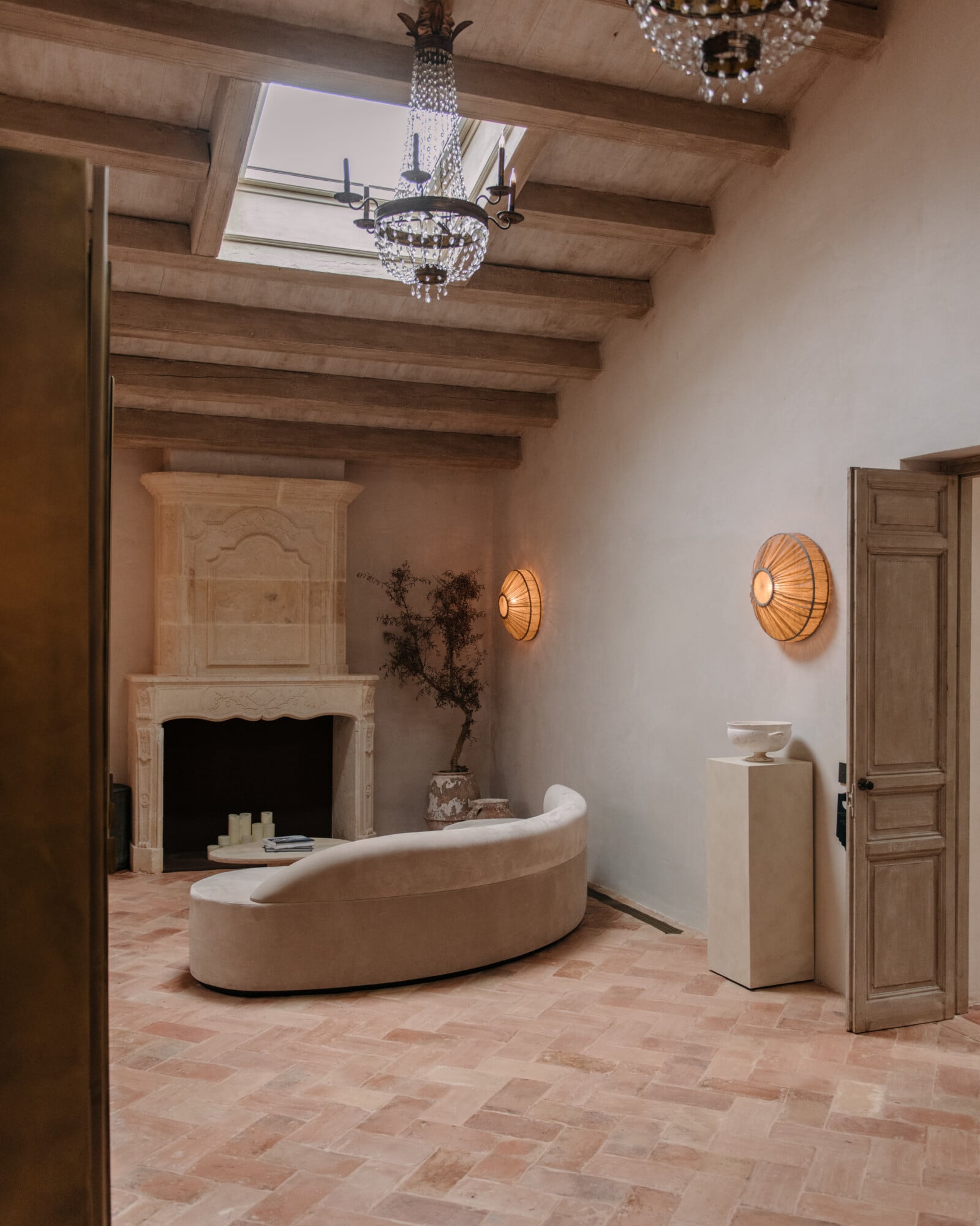 Son Vell, Balearic Islands, Spain — Curved cream sofa by fireplace under exposed wooden beams, terra-cotta tile floors, crystal chandeliers, minimalist luxury spa interior.