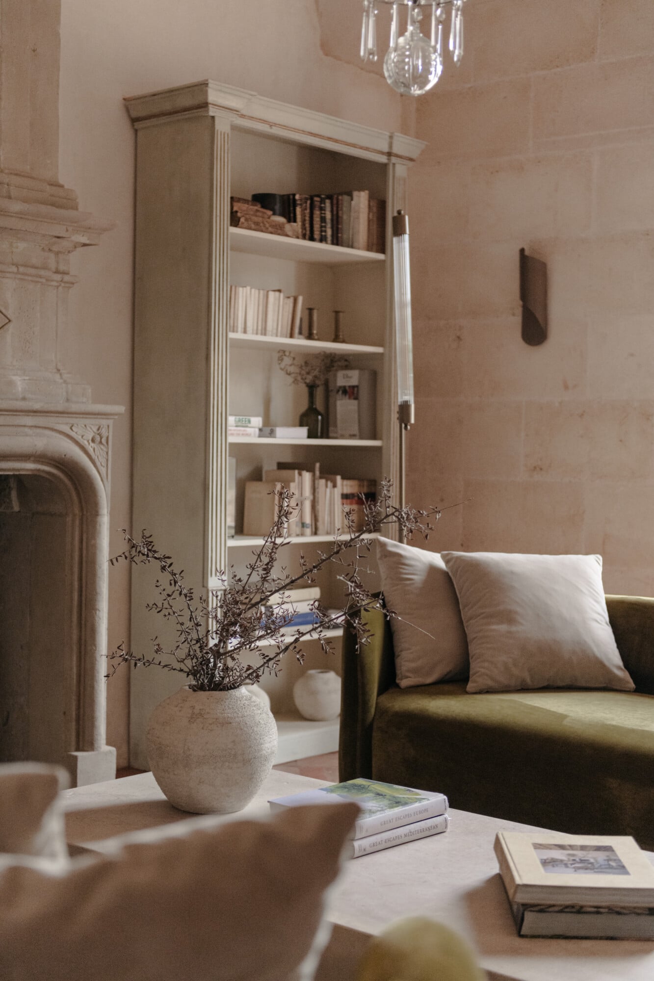 Son Vell, Balearic Islands, Spain — Cream-toned living room with fluted-trim bookcase, olive sofa, dried botanicals in textured vase, and crystal pendant light.