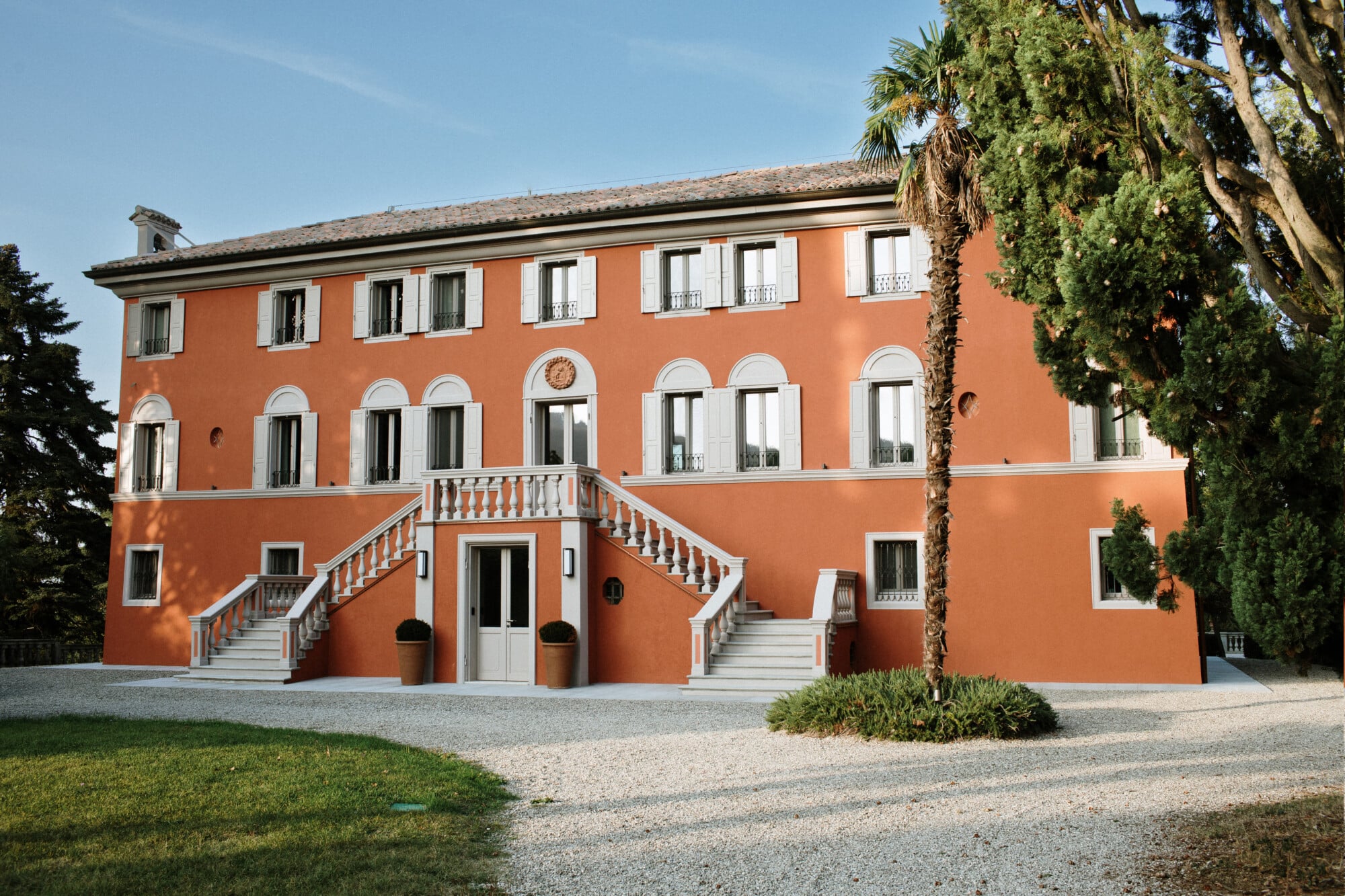 Roncolo 1888, Europe — Terracotta neoclassical villa with symmetrical arched windows, stone balustrade stairs, and lush green landscape under clear sky.
