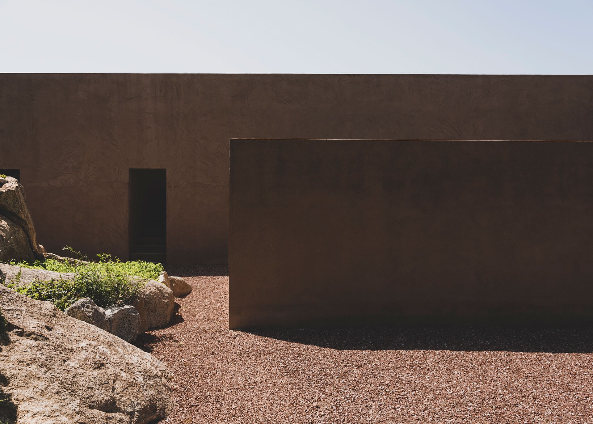 Villa Ra | MORQ — Minimalist brutalist courtyard with rammed-earth walls, gravel ground, natural rock elements, and dramatic shadow play under clear sky.