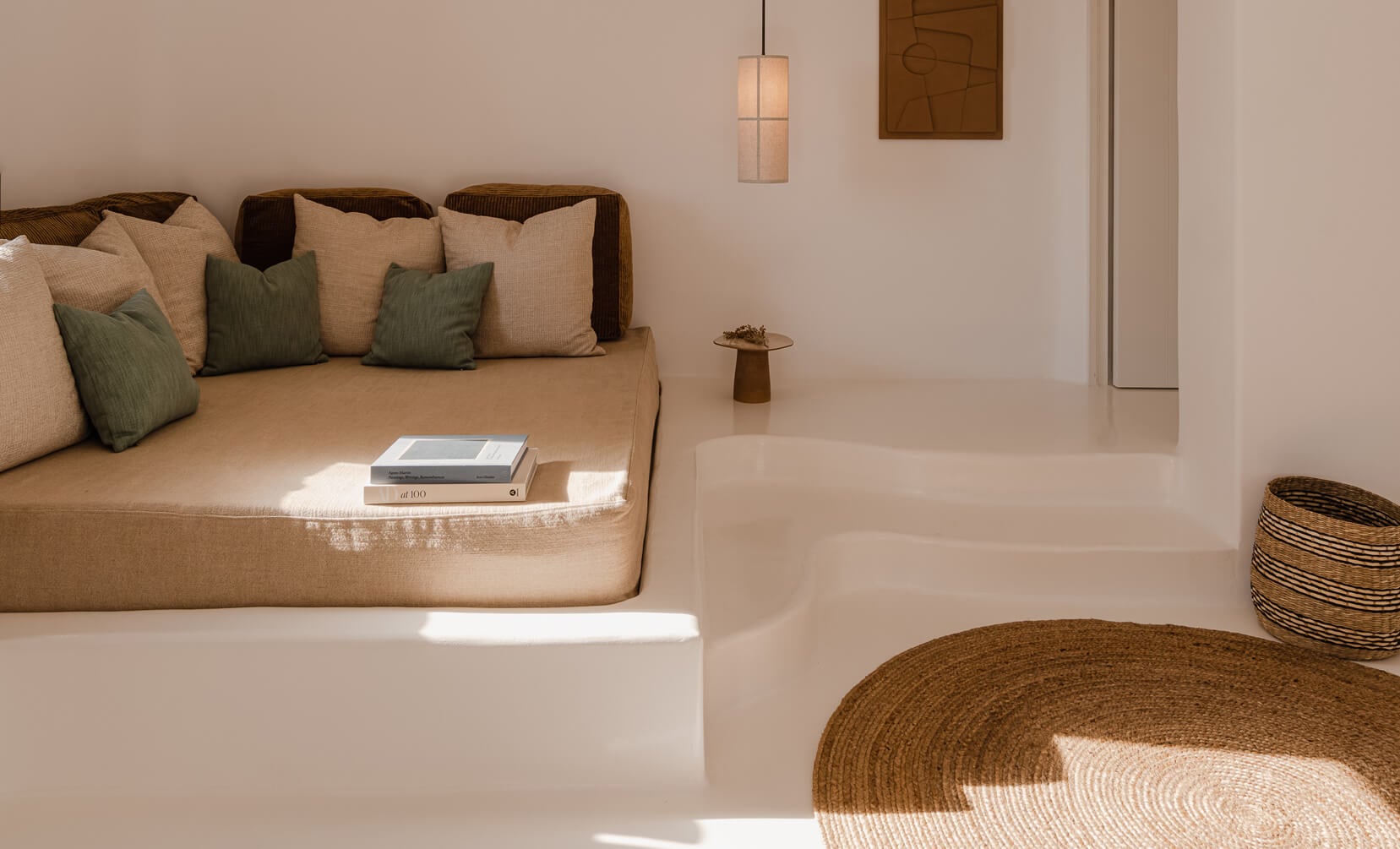 Nobu Santorini, Europe — Minimalist living room with beige linen sofa, sage pillows, natural light, woven round rug, pendant lamp, and warm neutral palette.