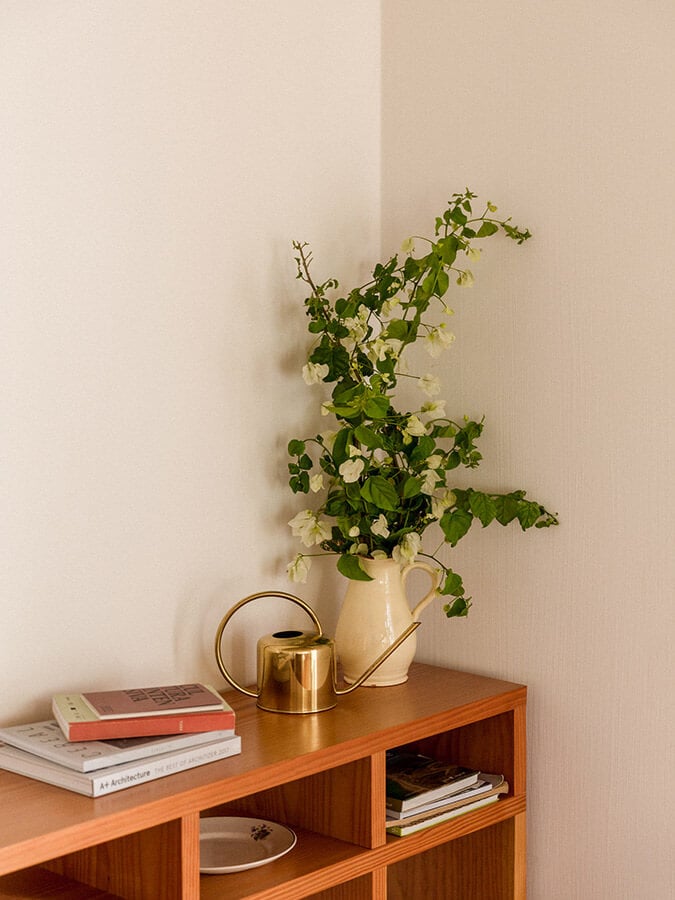 Casa Modesta, Europe — Wooden shelving unit with cream pitcher holding green foliage, brass watering can, and stacked books against neutral wall.
