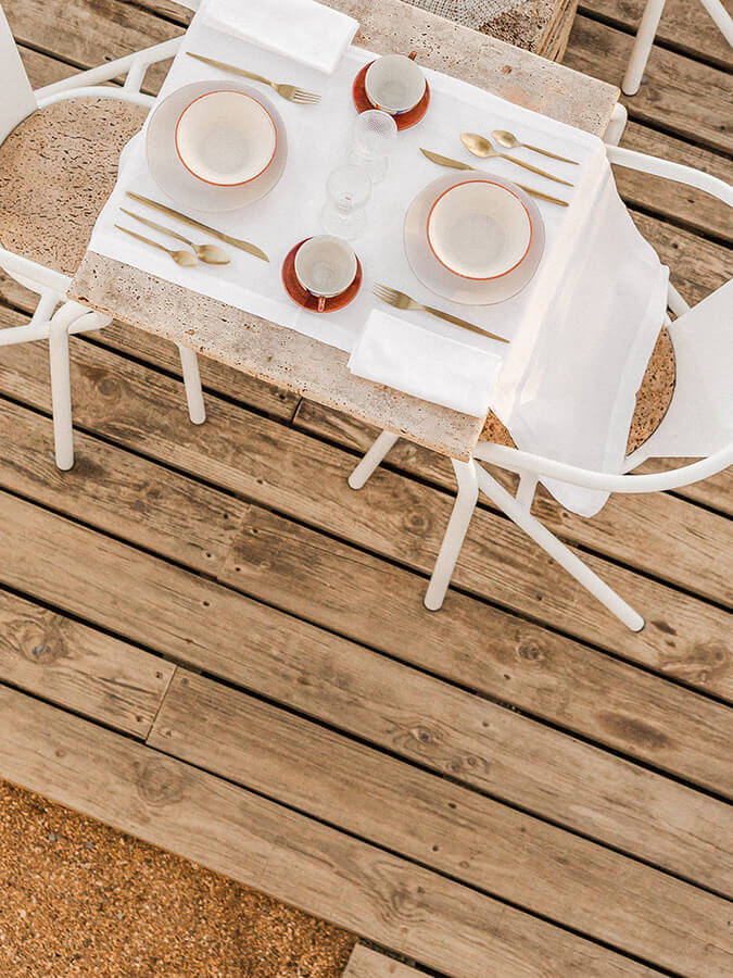 Casa Modesta, Europe — Overhead view of minimalist outdoor dining setup with white chairs, coral-rimmed plates, gold cutlery, and natural wood deck.