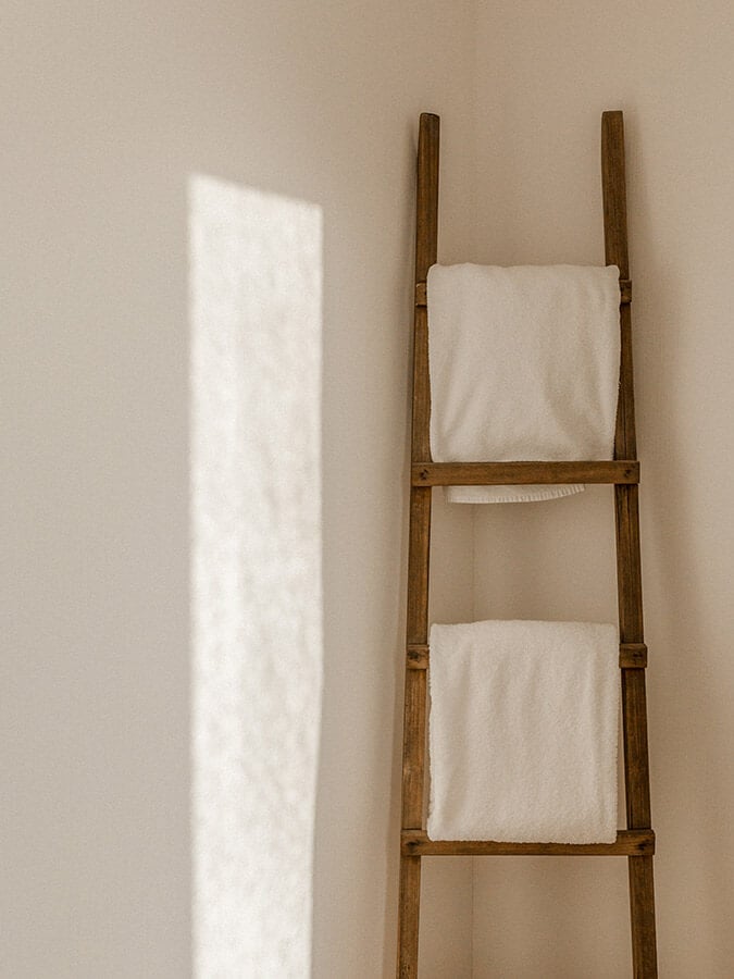 Casa Modesta, Europe — Wooden ladder with folded white linens against cream wall, natural sunlight casting geometric shadow, minimalist interior styling.