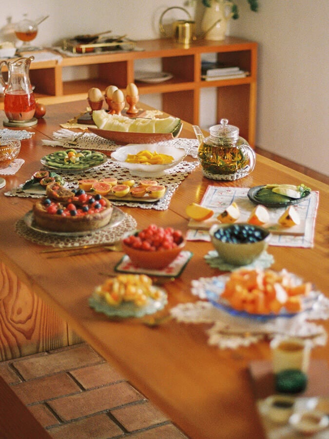 Casa Modesta, Europe — Wooden dining table styled with fresh fruits, vegetables, and appetizers arranged on white doilies, warm natural light, mid-century modern interior design.
