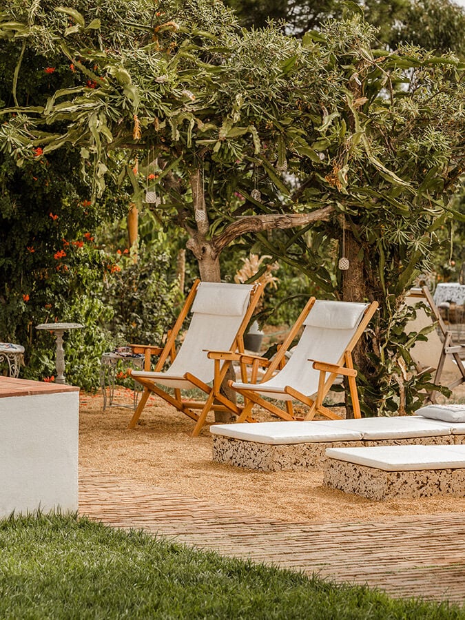 Casa Modesta, Europe — Wooden deck with teak lounge chairs and cream cushions beneath pergola draped in climbing vines, Mediterranean garden setting.