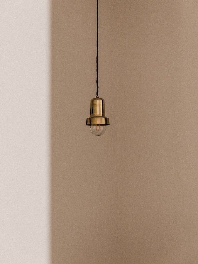 Casa Modesta, Europe — Brass pendant light with clear glass bulb suspended from black cord against warm taupe wall, minimalist hospitality lighting design.