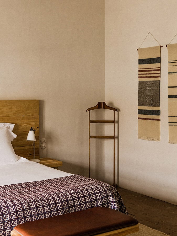 São Lourenço do Barrocal, Portugal — Minimalist bedroom with wooden headboard, striped wool wall hangings, metal valet stand, and terracotta tile flooring in warm neutral tones.
