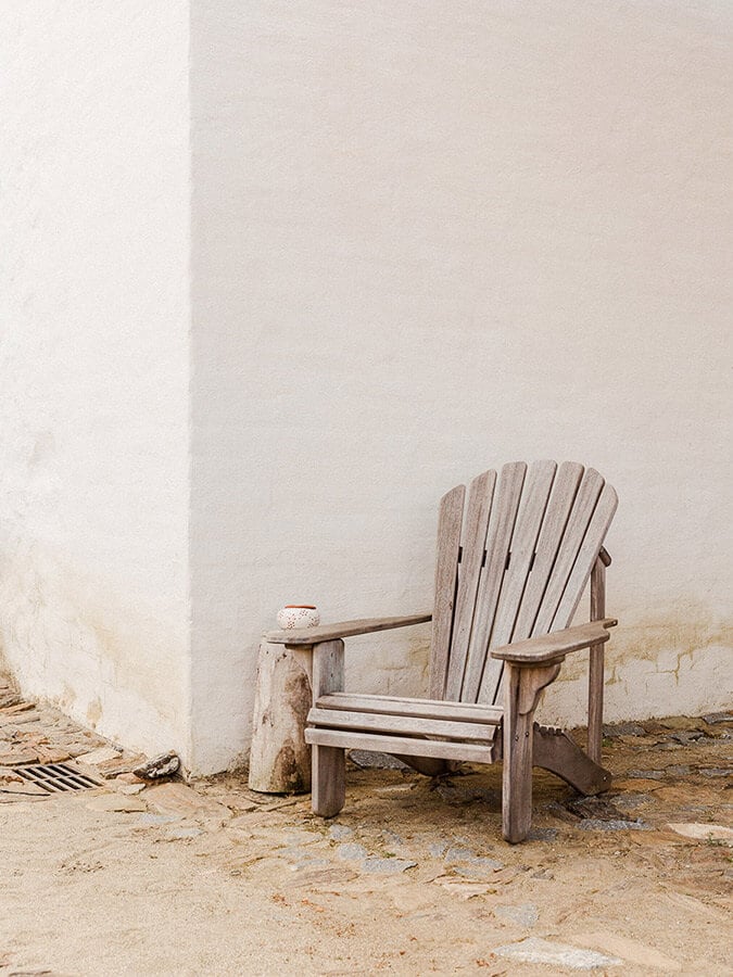 São Lourenço do Barrocal, Portugal — weathered wooden Adirondack chair against whitewashed stone wall on sandy ground, minimalist coastal aesthetic.