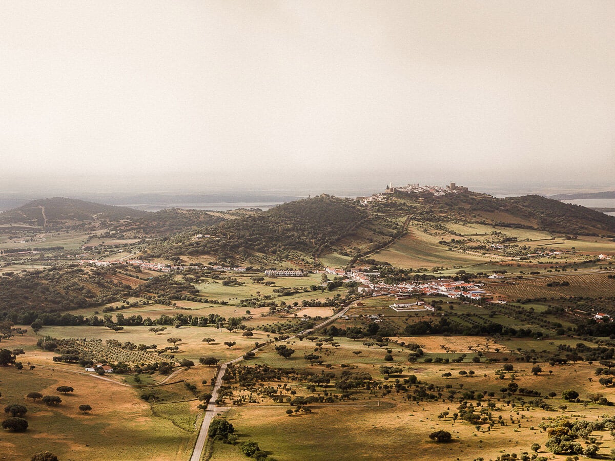 São Lourenço do Barrocal, Portugal — Aerial view of rolling countryside with whitewashed hilltop village, olive groves, and scattered cork oaks under hazy light.