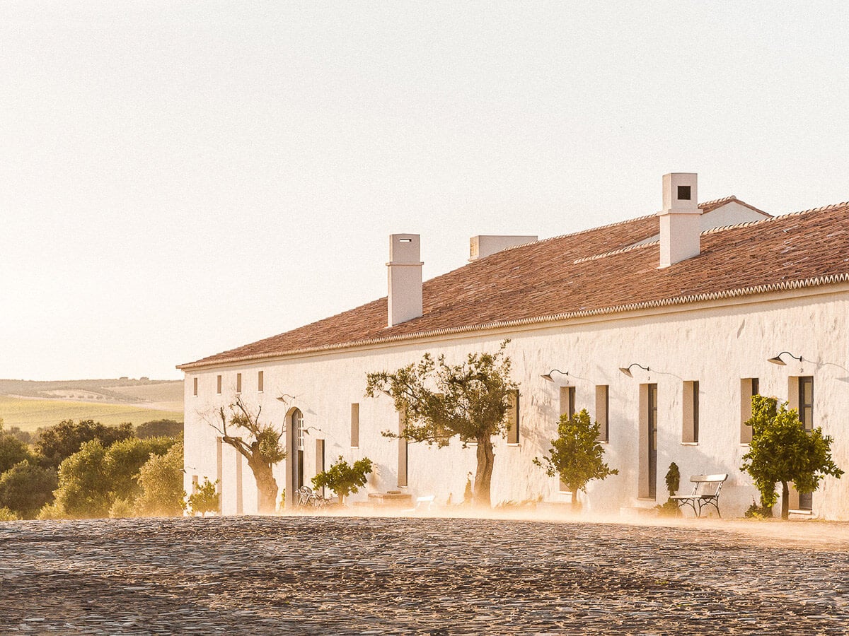 São Lourenço do Barrocal, Portugal — whitewashed rural estate with terracotta roof, olive trees, and golden-hour light casting long shadows across dusty courtyard.