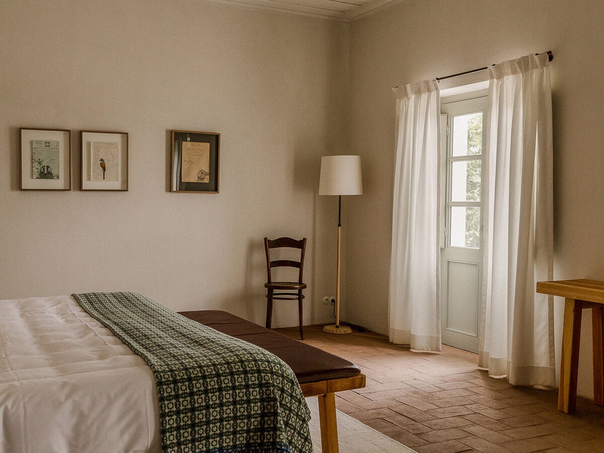 São Lourenço do Barrocal, Portugal — Minimalist bedroom with terracotta floors, cream walls, wooden furniture, and sheer curtains filtering natural light through tall windows.