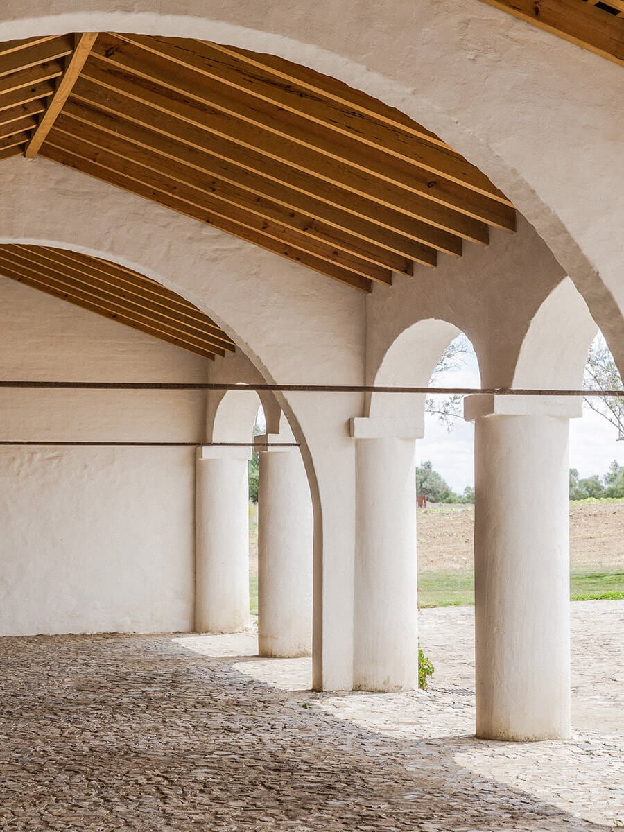 São Lourenço do Barrocal, Portugal — Whitewashed arched colonnade with cylindrical columns, wooden beam ceiling, and earth courtyard opening to rural landscape.