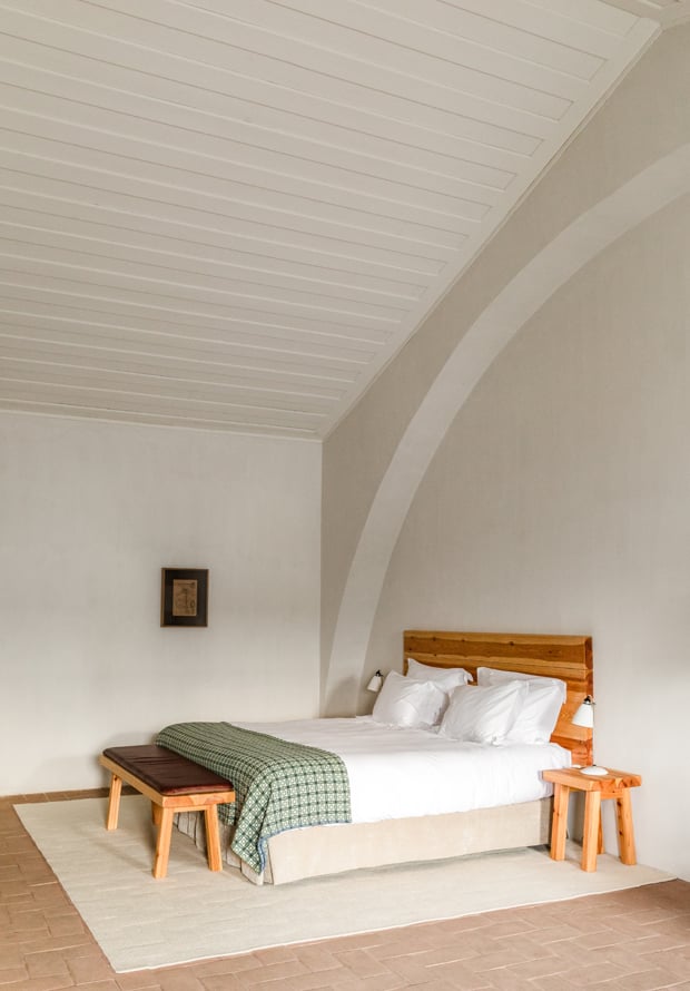 São Lourenço do Barrocal, Portugal — Minimalist bedroom with wooden bed, green throw, arched ceiling, white walls, terracotta floor, and natural light.