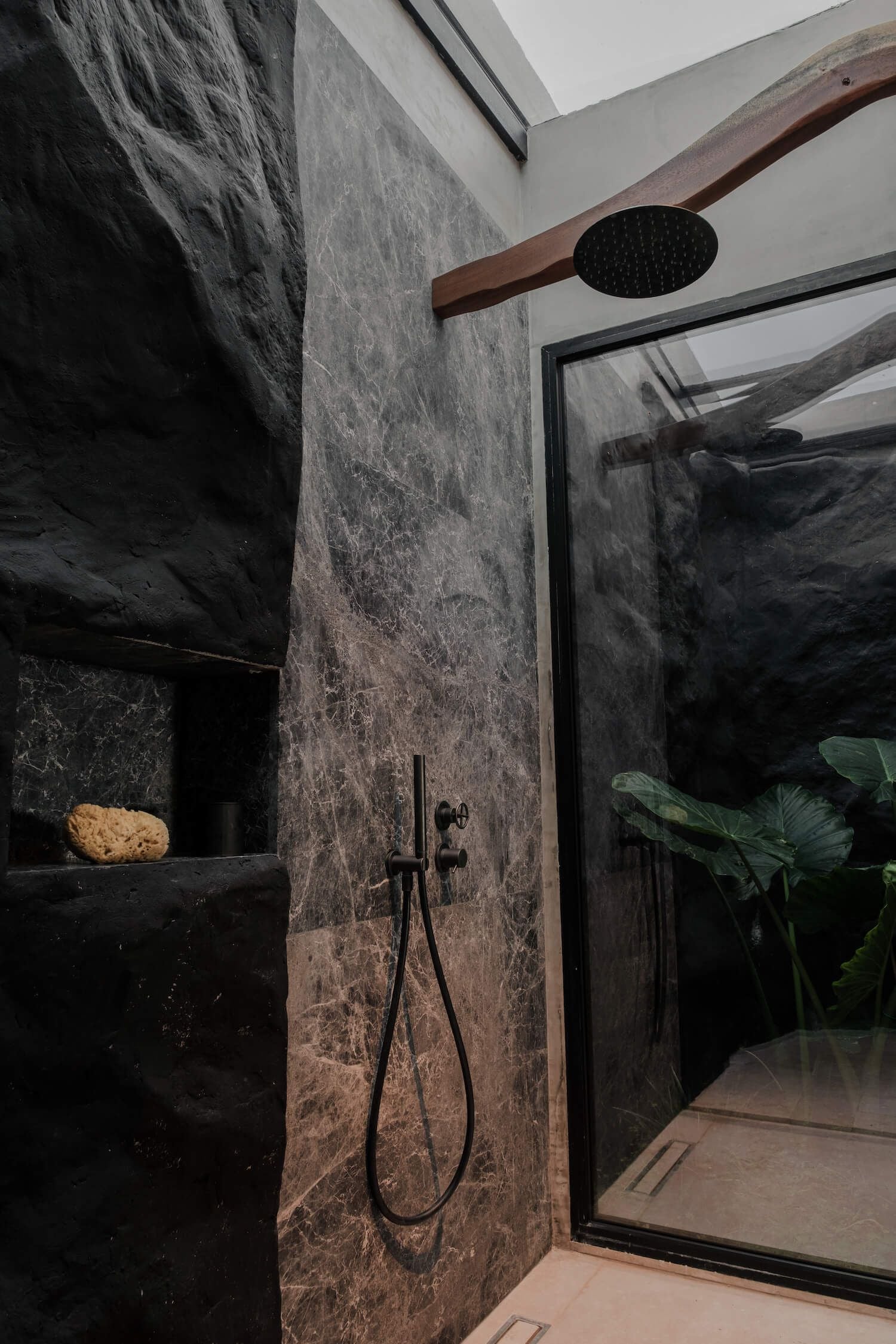 ACRO Suites, Europe — Rainfall showerhead suspended from timber beam above charcoal marble walls, black fixtures, and framed glass opening to lush greenery.