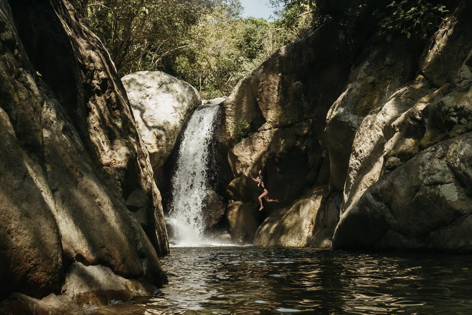 Sierra Madre, Mexico — Natural waterfall cascading through limestone canyon with emerald pool, moss-covered rock formations, and dappled forest canopy light.