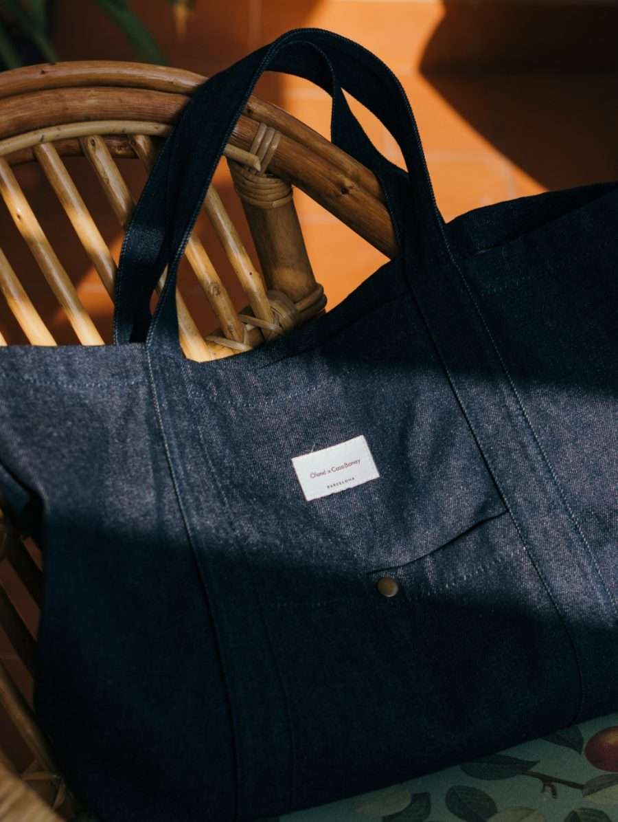 Casa Bonay, Europe — Dark denim tote bag with leather handles draped over wooden chair, warm golden light, minimalist luxury aesthetic.
