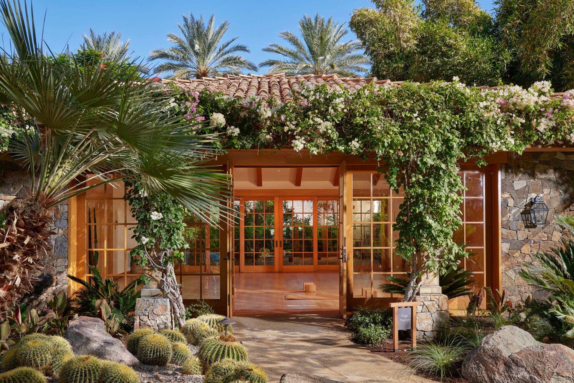 Meditation & Mindfulness, North America — Wooden pergola with flowing white flowers, stone walls, palm trees, and desert landscaping creating serene courtyard entry.