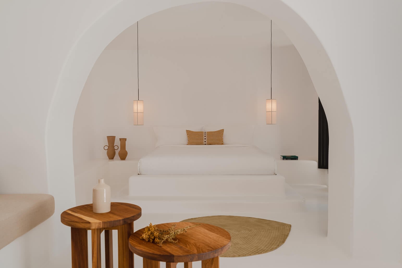 Nobu Santorini, Greece — Minimalist whitewashed bedroom with arched alcove, floating bed, pendant lights, wooden side tables, and warm neutral accents.
