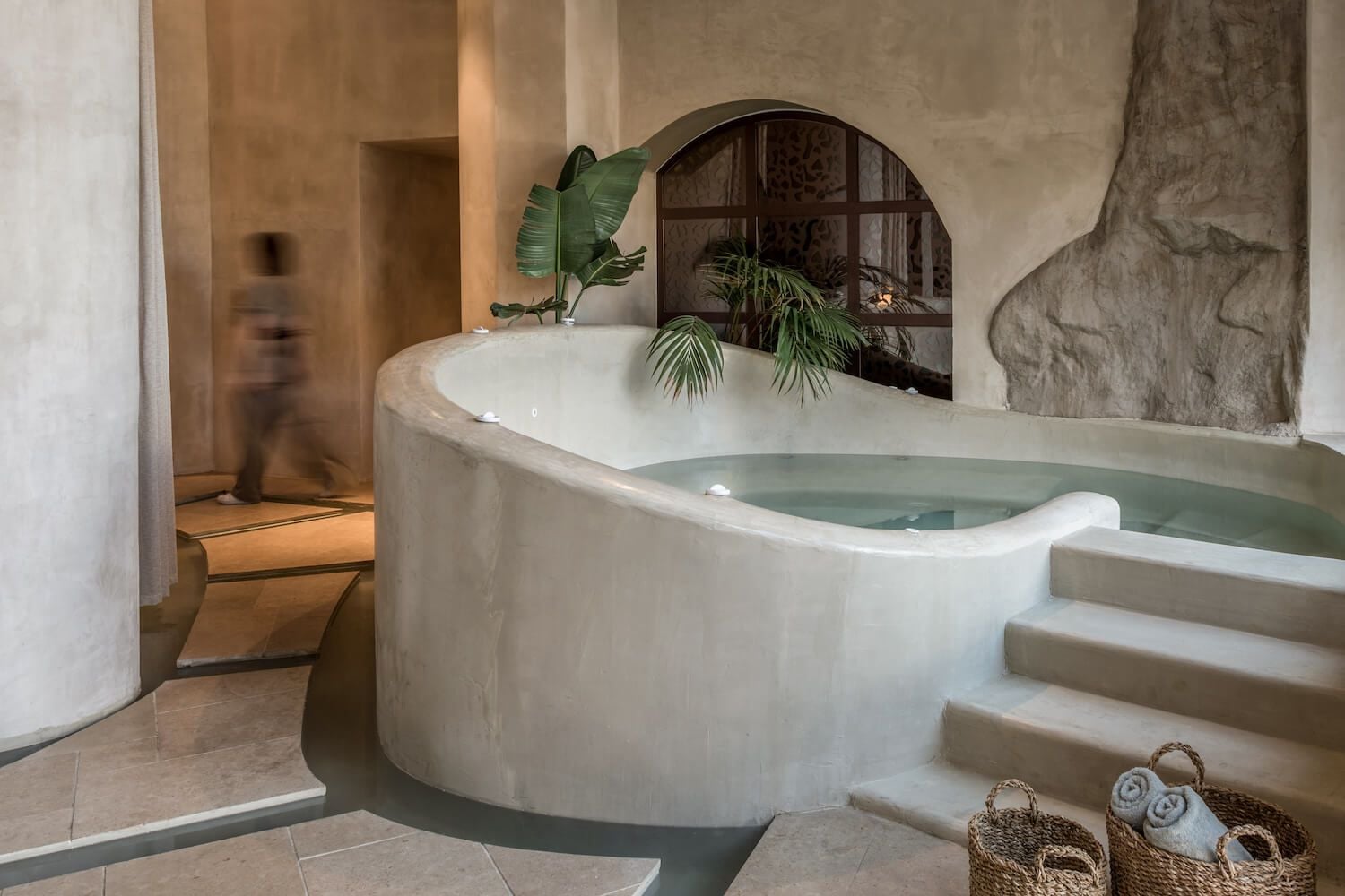 The Byzantine Hammam, Europe — Sunken marble bath with stepping stones, arched mirror, potted plants, and warm stone architecture bathed in soft natural light.