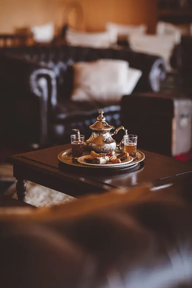 Scarabeo Camp, Africa — Brass Moroccan teapot with glasses and pastries on gold tray, dark wood table, warm ambient lighting, luxury lounge interior.