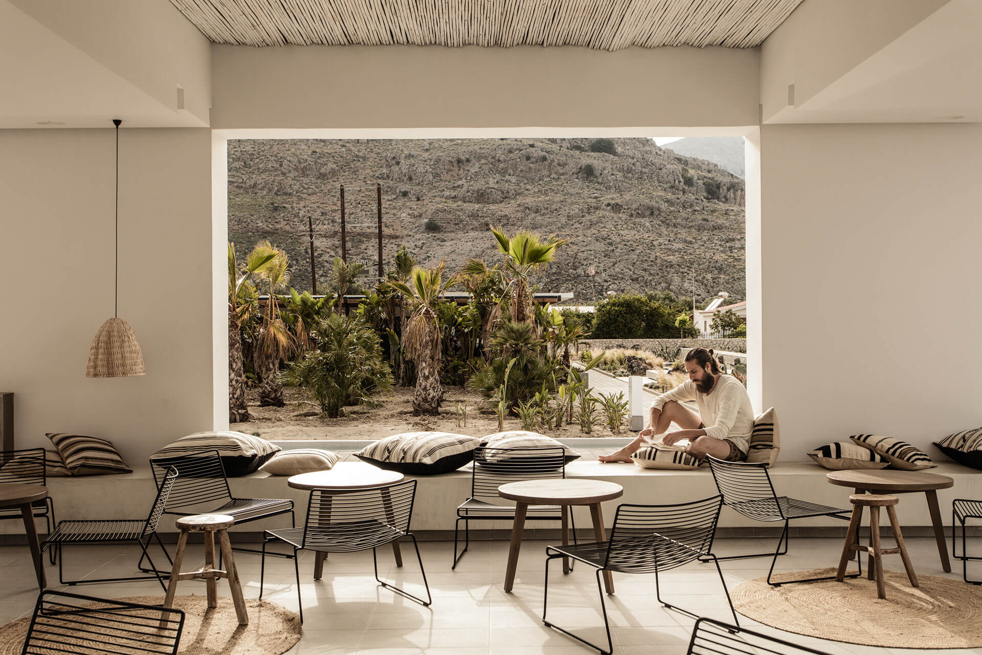 Casa Cook Rhodes, Greece — Minimalist lounge with striped chairs, cushioned seating, woven pendant light, and expansive view of desert mountain landscape with palm trees.