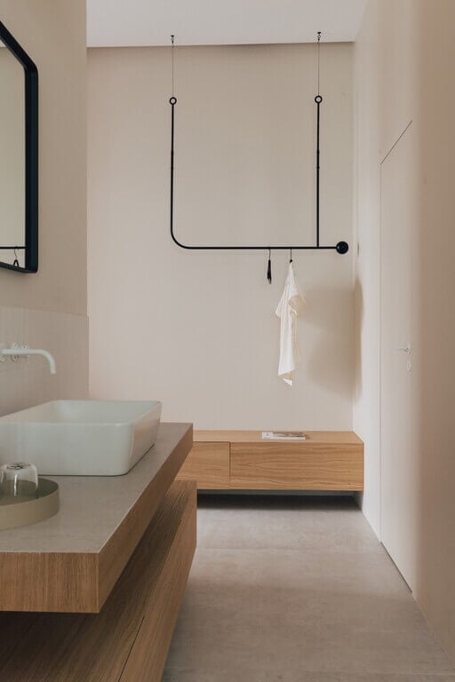 Plácido y Grata, Europe — Minimalist bathroom with suspended black pipe towel rail, natural wood vanity, pale stone walls, and concrete flooring.