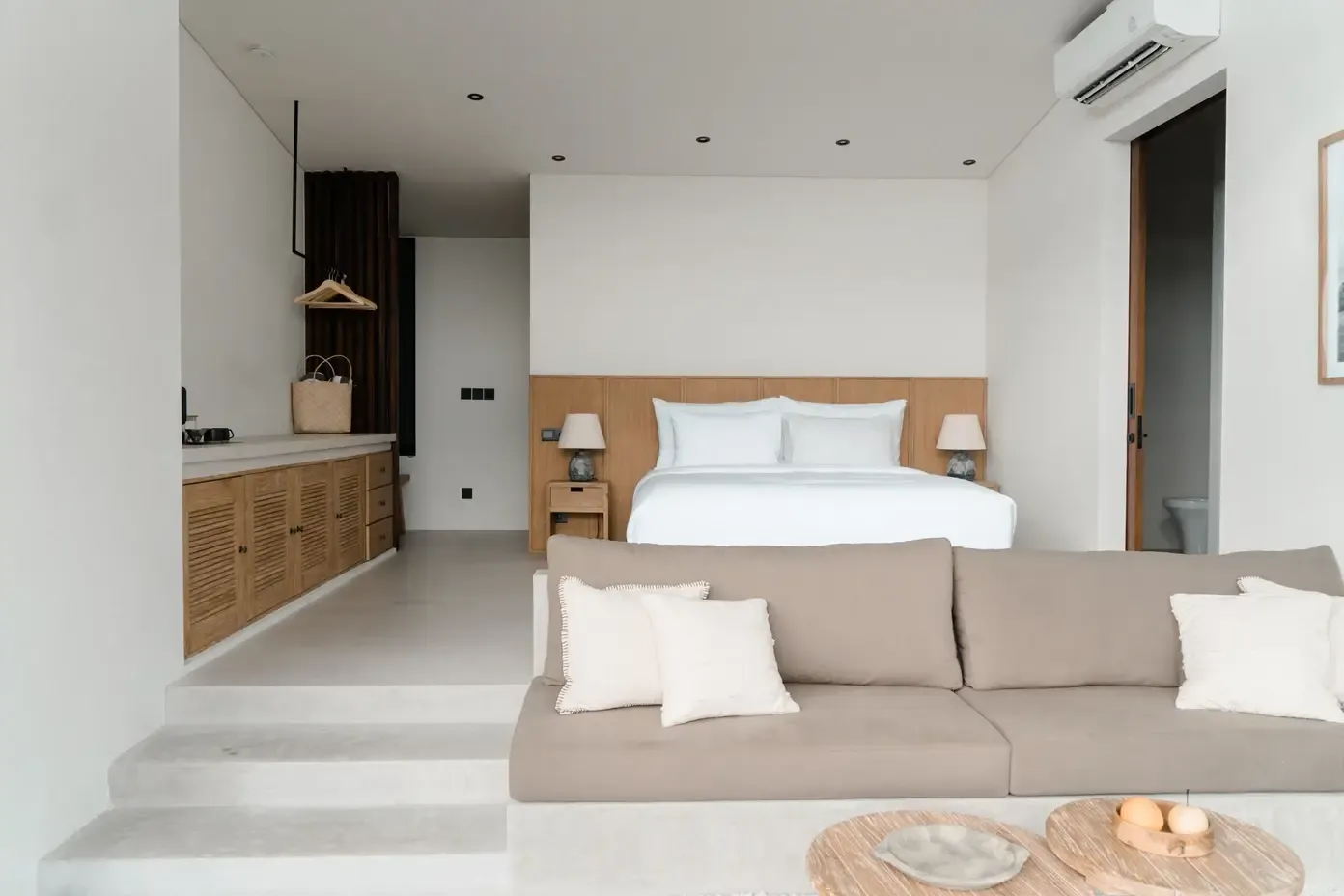 Somewhere Lombok, Asia — Modern studio bedroom with wood-paneled headwall, beige linen sofa, white bedding, natural materials, and minimalist neutral interior design.