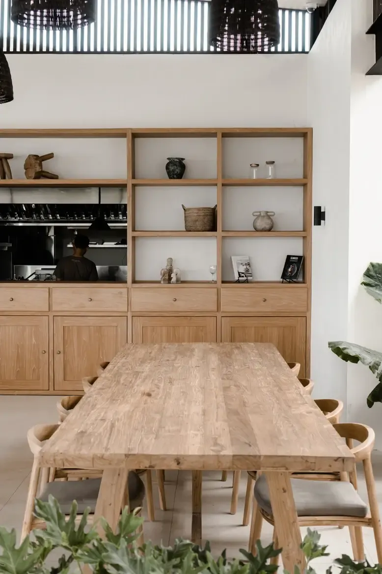 Somewhere Lombok, Asia — Light oak dining table and chairs with sculptural cutouts facing built-in shelving unit displaying ceramics and decorative objects in minimalist interior.