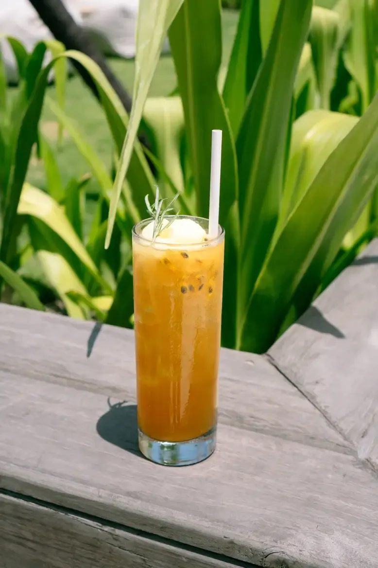 Somewhere Lombok, Asia — Amber cocktail with rosemary sprig and ice on weathered wood table, lush green plants behind, tropical daylight.