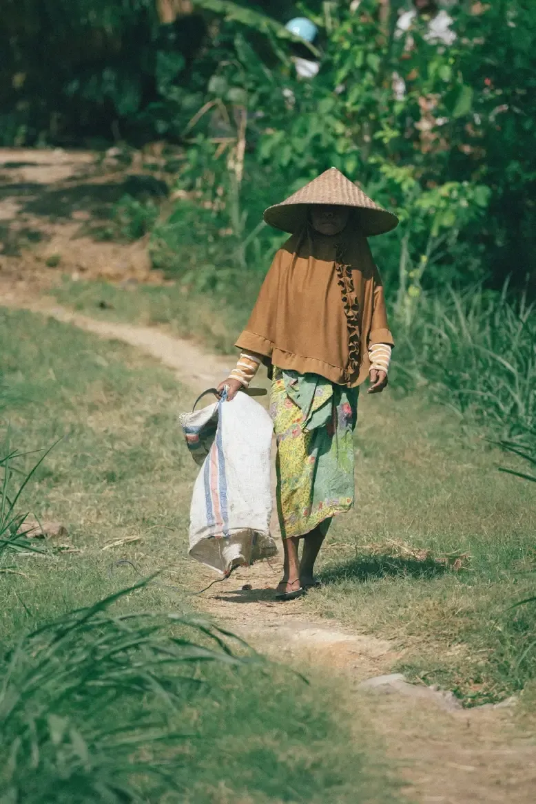 Somewhere Lombok, Asia — Woman in conical hat walking rural path through lush greenery, wearing traditional ochre tunic and patterned sarong, carrying white fabric bundle.