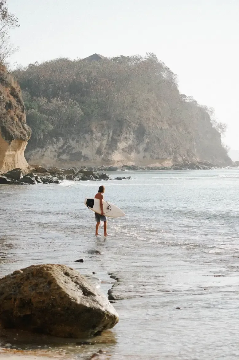 Somewhere Lombok, Indonesia — Surfer in ocean with limestone cliff and forested headland, golden hour coastal light.